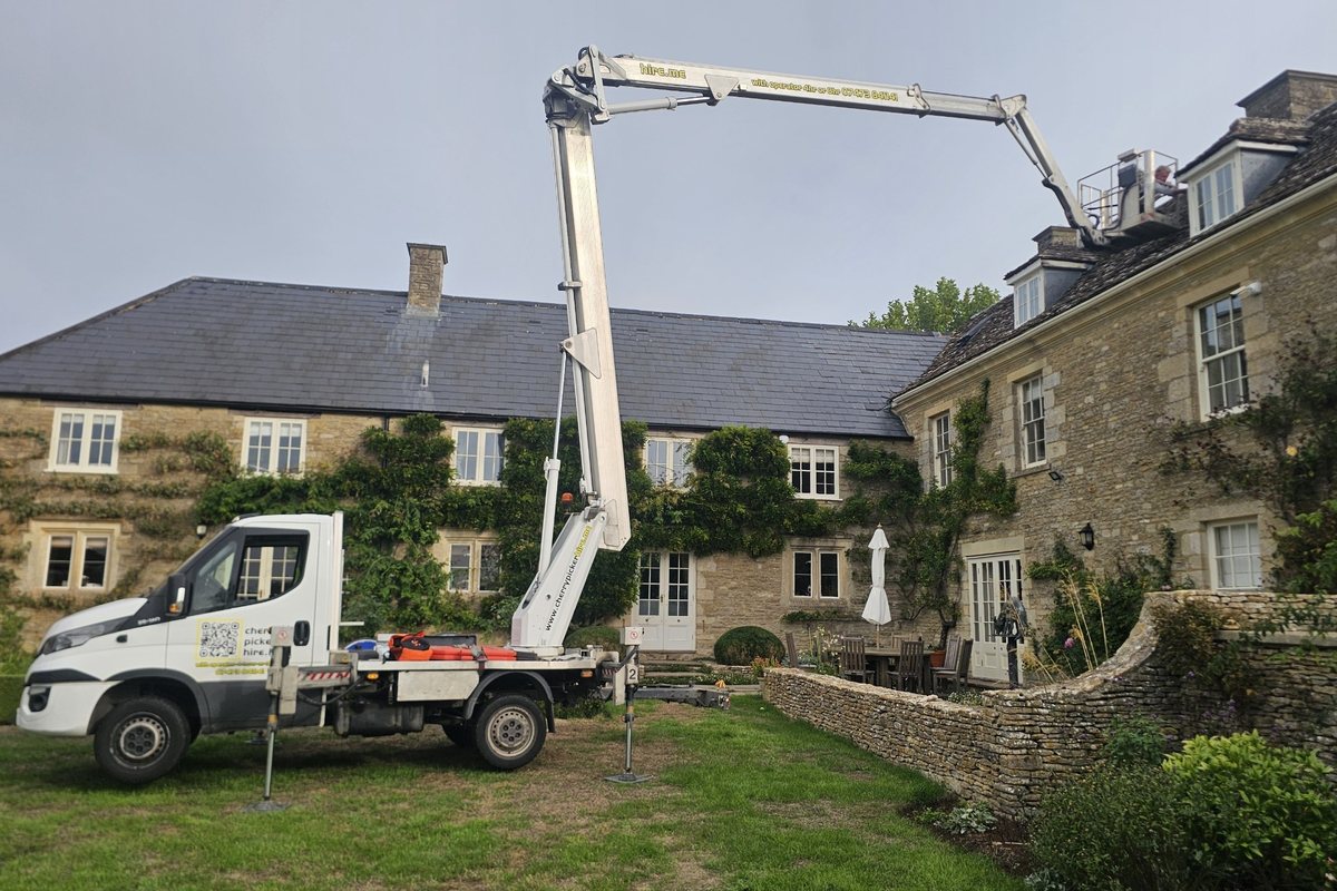 Cherry picker set up on the lawn of a Cotswold stone manor house with the boom reaching over the slate roof to a dormer window