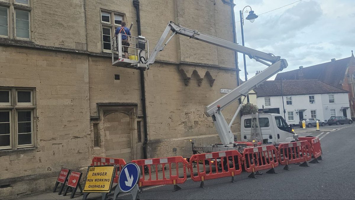 Operator working at first-floor level on a historic stone building in a town centre, with pedestrian barriers and overhead-working warning signs in place